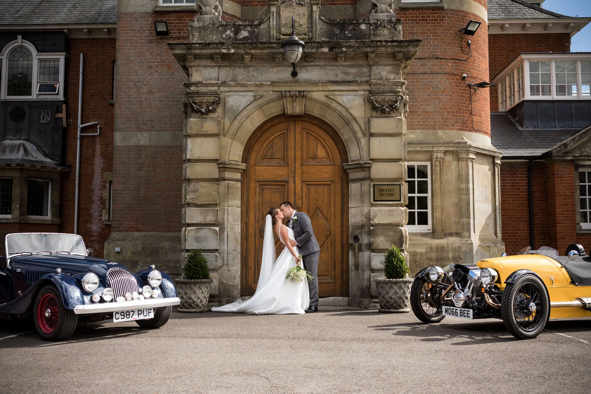 Berkshire wedding photographer - couple kiss outside Pangbourne College with classic cars on either side of the doorway used for the background.