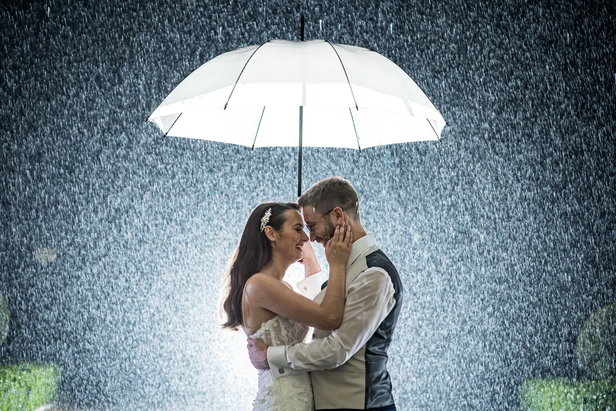 Sussex wedding photographer - a couple laugh together under a umbrella as a deluge of rain falls in the background, lit magically by external lighting