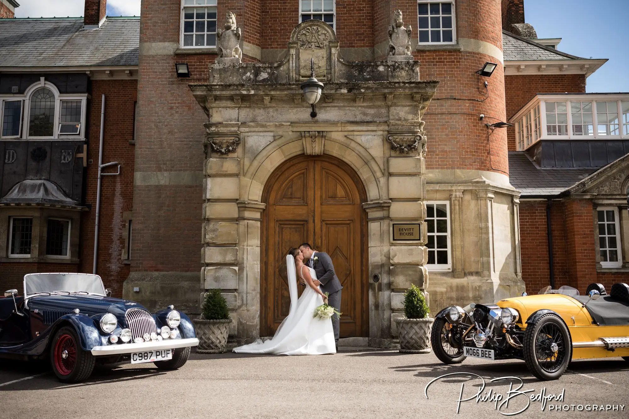 Bride and groom kissing in front of a dark doorway with two Morgan cars on either side.